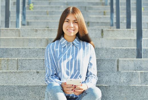 A young girl of Asian appearance, a student dressed in jeans and a striped shirt, holds a tablet in her hands and sits on the stairs smiling at the camera