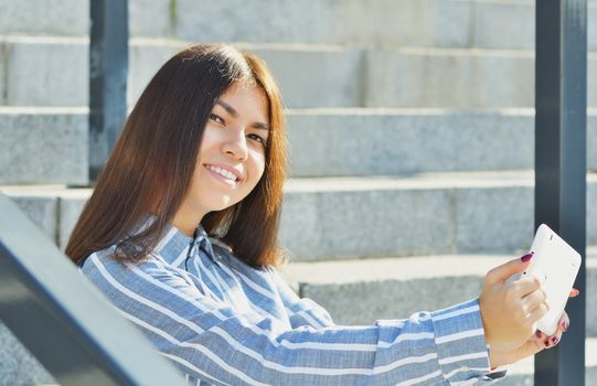 Young beautiful girl student Asian appearance, holding a tablet and talking on video, sitting on the steps in the street, smiling at the camera