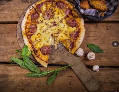 Delicious fresh made pizza made with pepperoni, mozarella, chilli served on a timber pizza board with fresh basil and mushrooms scattered around on a rustic timber table