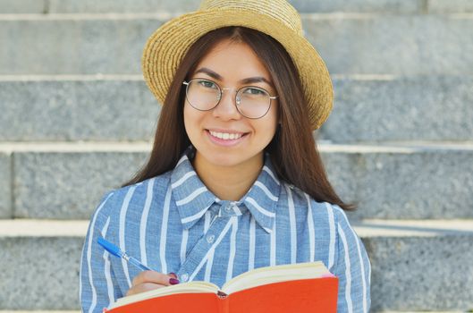 Portrait of a young Asian student in glasses with a hat dressed in a blue striped shirt who writes tasks in a notebook, and smiles at the camera