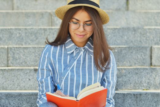 Portrait of a young Asian student in glasses with a hat dressed in a blue striped shirt that looks at the notebook writing tasks