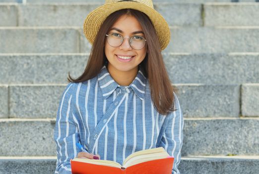 Portrait of a young Asian student in glasses with a hat dressed in a blue striped shirt who writes tasks in a notebook, and smiles at the camera