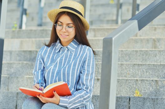 Portrait of a young Asian student in glasses with a hat dressed in a blue striped shirt that holds a notebook and writes tasks