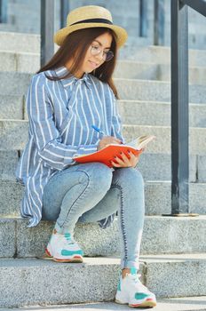Portrait of a young Asian student in glasses with a hat dressed in a blue striped shirt and sneakers, the girl is holding a notebook and writing tasks