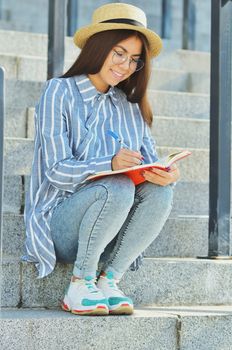 Portrait of a young Asian student in glasses with a hat dressed in a blue striped shirt and sneakers, the girl is holding a notebook and writing tasks