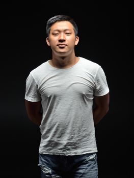 Portrait of a male Asian student with a black background, who put his hands behind his back and looks straight