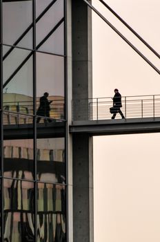 Businessman walking across bridge towards business building, nobodz, reflection in glass facade, holding a mobile phone, business and executive concept