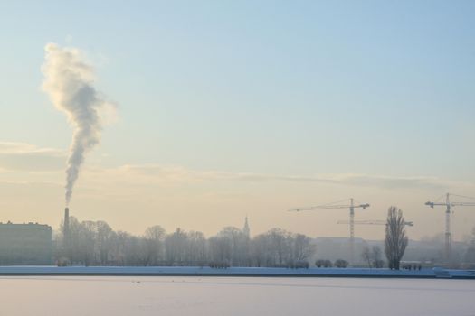 Riga view in winter; Riga, capital city of Latvia in winter time. View of Riga with frozen river Daugava and smoky chimney. City Riga with background of blue sky. 