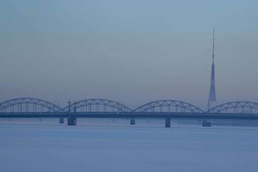 Railway bridge and television tower in winter. Railway bridge over the frozen river Daugava in Riga.TV tower and railway bridge on blue sky background in winter. Riga view in winter.