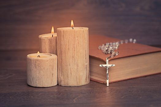 Silver rosary and crucifix resting on closed book near the candles on wooden table, religion school concept. Vintage style.