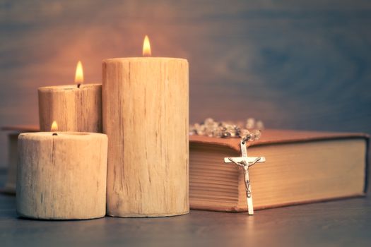 Silver rosary and crucifix resting on closed book near the candles on wooden table, religion school concept. Vintage style.