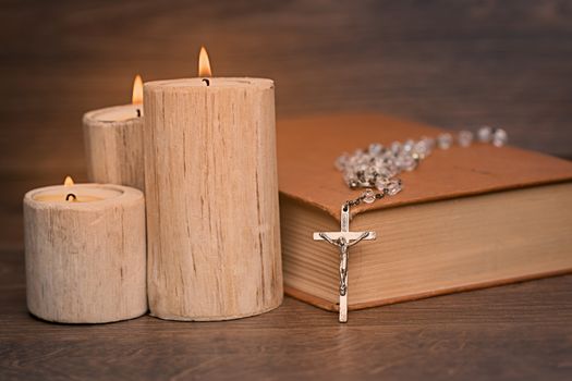 Silver rosary and crucifix resting on closed book near the candles on wooden table, religion school concept. Vintage style.