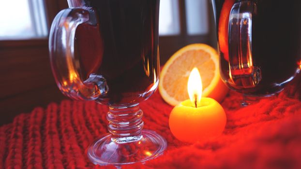 Mulled wine in glass mugs, burning candle on a dark red background. Red Hot wine with orange and red scarf