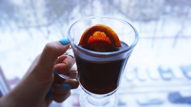 Woman holding mulled wine with spices and citrus fruit in her hand, close up in front of the window with a winter view