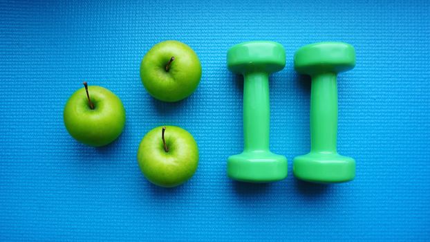 Dumbbells near green apples on blue background. Healthy lifestyle and sports concept. Apple fruit and green barbells. Health regime and fitness symbols