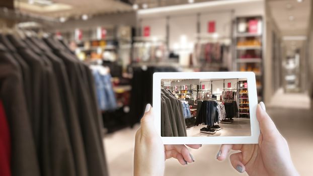 Woman hands taking a tablet in shopping mall with blurred image of clothes shop. The concept of online shopping and augmented reality