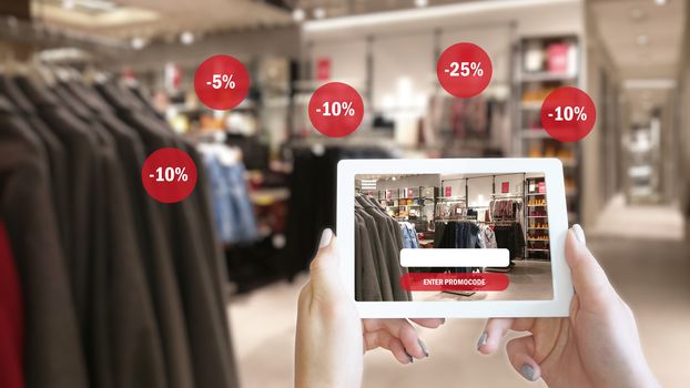 Woman hands taking a tablet in shopping mall with blurred image of clothes shop. The concept of online shopping and augmented reality - Enter promo code to get a discount online