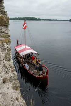 Boat at Koknese castle ruins. Tourist boat in the style of viking on river Daugava, Latvia. Tourists in old wooden viking boat, next to the castle Koknese, Latvia. The medieval castle of Koknese was a stone castle built in the medieval castle mound in Koknese, on the bank of the Daugava. Built shortly after 1209.