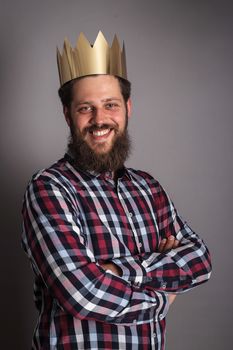 Bearded man in golden crown looking at camera while standing with crossed arms on gray background