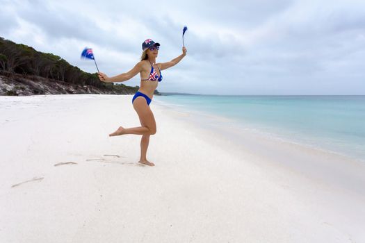 Patriotic female on the beach with Australian flag on  her hat, sunglasses and bikini and she is also waving two small flags while enjoying the idyllic beaches of Australia
