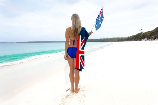 Woman standing on white sandy beach, she is waving a flag and has an Australian flag towel draped over her shoulder