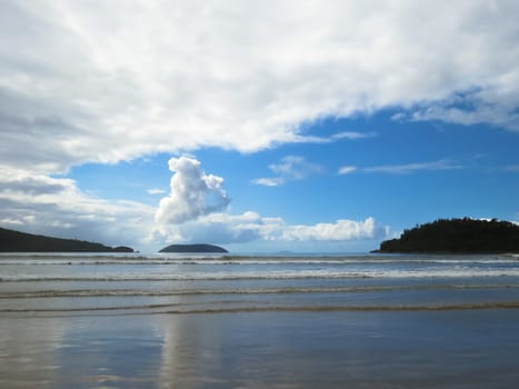 Beach with small waves in blue sky day and clouds, with islands on the horizon.