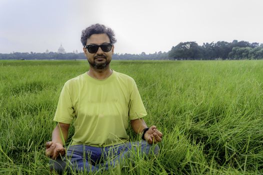 Yoga at park. Senior bearded man in lotus pose sitting on green grass. Concept of calm and meditation.Young man in sunglasses meditating outdoors in the park. ( Moidan, Kolkata, India)
