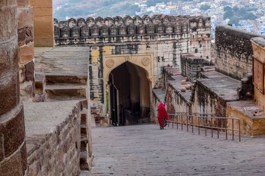 Portrait of a poor rural indian lady in traditional red saree or sari walking one morning summer in UNESCO architectural heritage building. ( Jaisalmir Fort, Jaipur City road, Rajasthan, India, Asia)