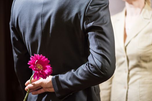 A young man in a suit holds a gerbera flower behind his back, a surprise for a woman, March 8.