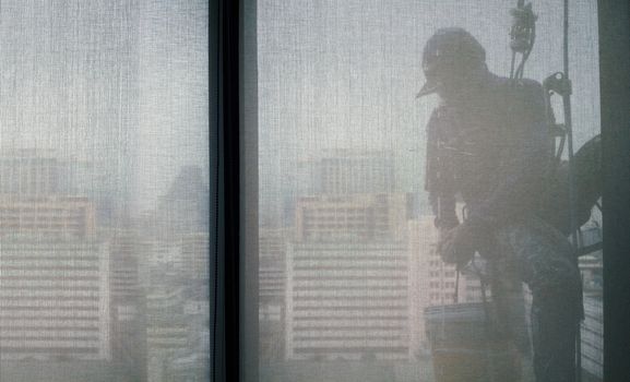 Silhouette images and shoot through transperent curtain effect from inside building which a man cleaning the window of high office building with his equipment such as wipe, sponge, bucket and high risk of danger