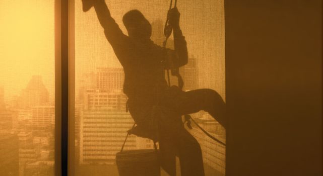 Silhouette images and shoot through transperent curtain effect from inside building which a man cleaning the window of high office building with his equipment such as wipe, sponge, bucket and high risk of danger