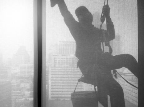 Silhouette images and shoot through transperent curtain effect from inside building which a man cleaning the window of high office building with his equipment such as wipe, sponge, bucket and high risk of danger