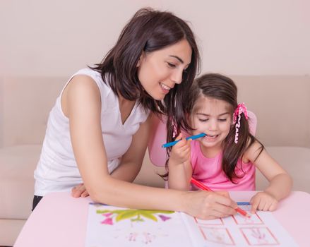 Mother and daughter study activity book with pencils on paper while sitting at pink table.Preschool and kindergarten education at home.Selective focus and small depth of field.