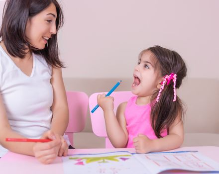 Mother and daughter study activity book with pencils on paper while sitting at pink table.Preschool and kindergarten education at home.Selective focus and small depth of field.