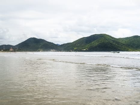 Sunny beach with small waves and bathers and hills covered with forest in the background.