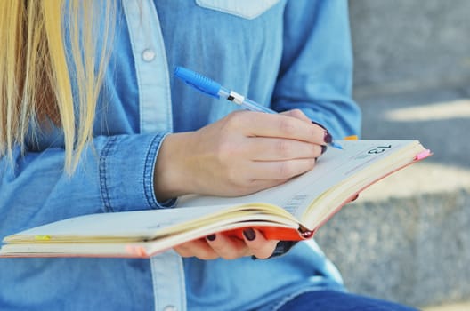 The hand of a young girl dressed in a denim shirt, who writes in a notebook on the street, sitting on the steps