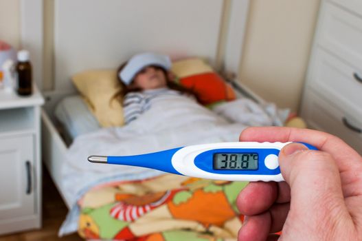 close up on a digital thermometer  that marks 38 degrees Celsius.  Little girl into bed with wet cloths on forehead