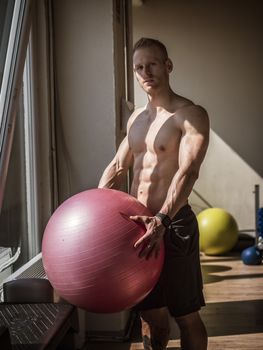Attractive blond young man shirtless in gym working out, holding inflatable fitness ball, about to do exercises for abs.