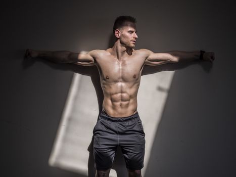 Attractive shirtless blond male bodybuilder in shorts indoors in dark gym, showing muscular torso and ripped abs