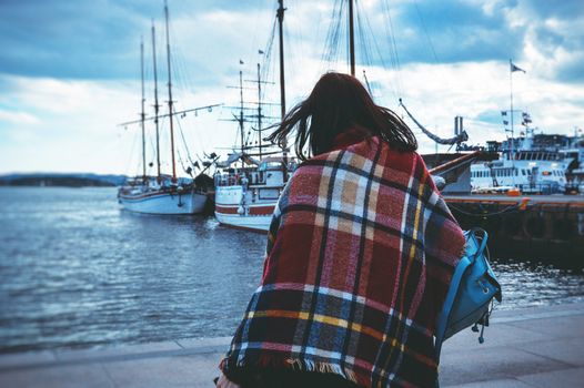 Young beautiful girl in a warm plaid walks in the port near boats in cold weather in Stockholm