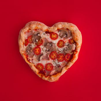 Pizza heart shaped with ham tomatoes and mushrooms on red background. Concept of romantic love for Valentines Day . Love food