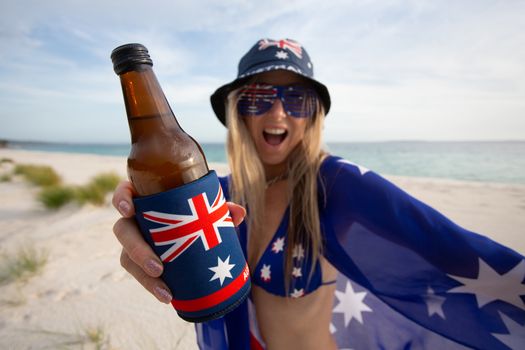 A revelling woman holding a beer on the beach celebrates Australia Day or supports Australian sport enthusiast fan. Beach culture or Aussie holiday.  Focus to bottle