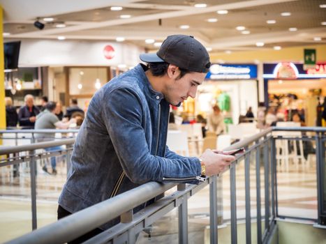 Young handsome trendy man in shopping mall, with cell phone in hand, typing or reading