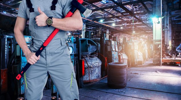 Closeup of worker man with ax dressed in overalls on Industrial zone background, manufacturing facility. Concept of Safe labor in the enterprise.