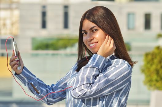 A young girl student of Asian appearance dressed in a striped shirt listens to music while on the street