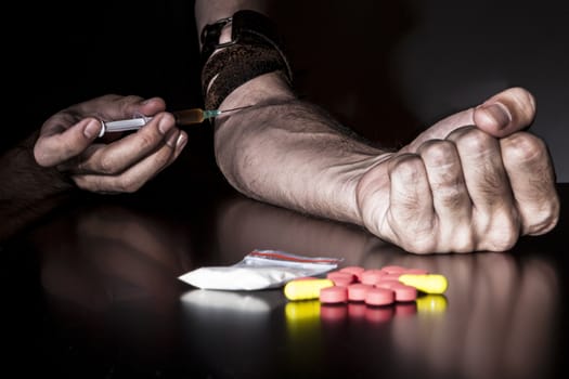 Injecting drugs with syringe with colored pills and bag of powder in the foreground