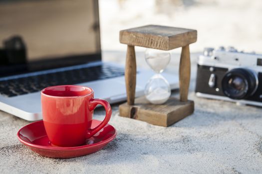 Red Coffe Mug and Vintage hourglass with silver laptop and old camera in the sand at the beach
