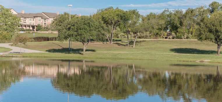 Panorama view multistory apartment complex near city park with pathway, lake and cloud sky reflection. Natural rental area near Dallas, Texas, USA