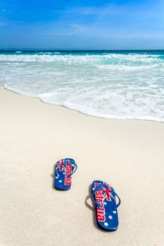 Australian flag printed on thongs on the beachwith an encrouching wave coming toward them  in summer sun