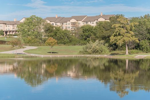 Multistory apartment complex near city park with pathway, lake and cloud sky reflection. Natural rental area near Dallas, Texas, USA